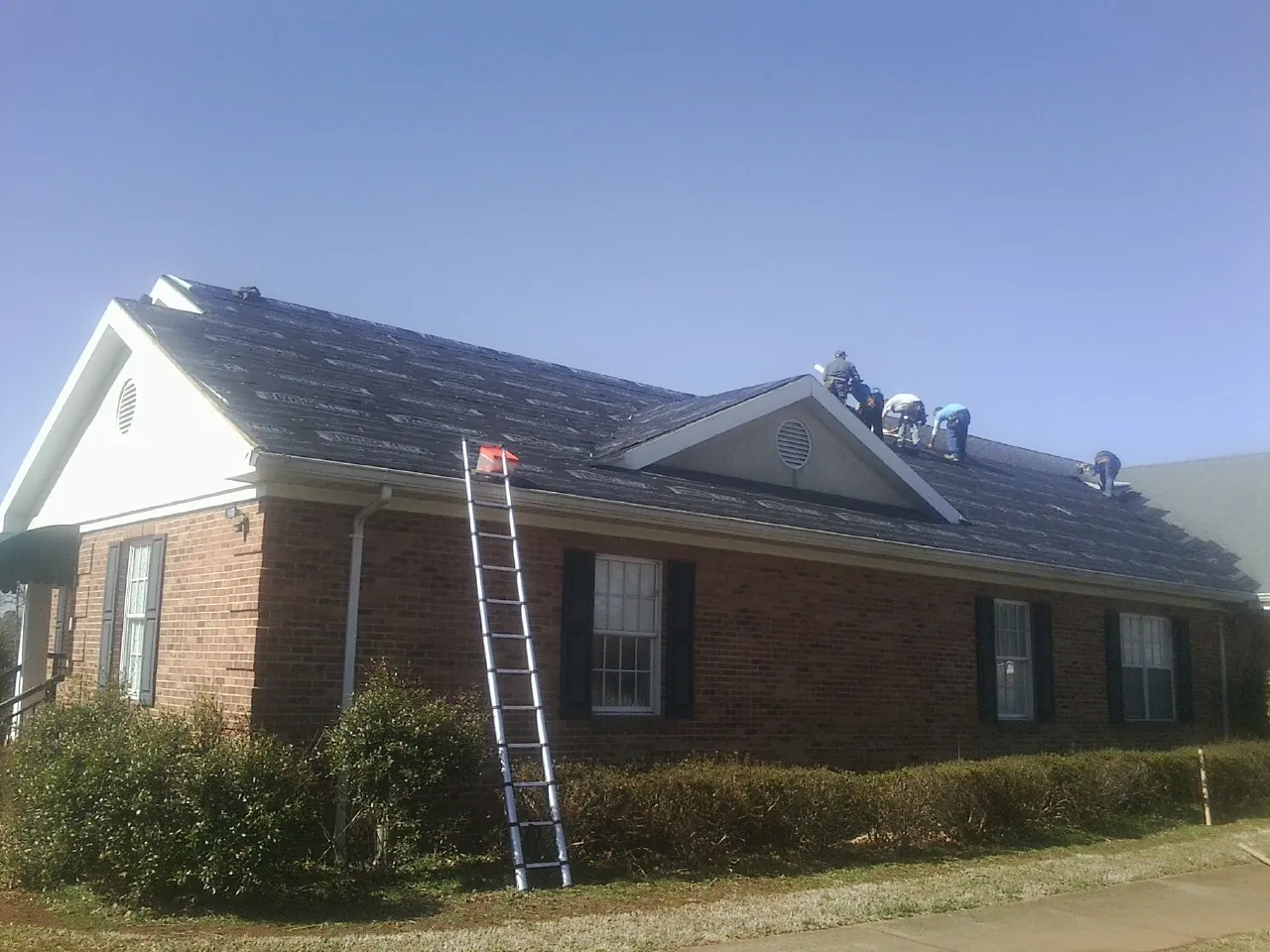 Crew tearing shingles off a brick ranch roof with a ladder set — Metal Roof Repair work by our crew in North Vernon