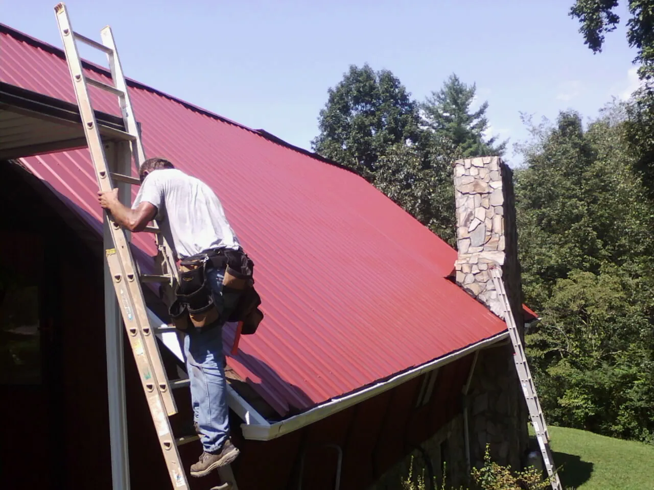 Roofer on a ladder beside a red metal roof and stone chimney — Soffit & Fascia Repair by Roofing Brothers in North Vernon